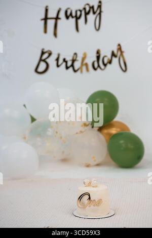 Charmante configuration de premier anniversaire avec une bannière joyeux anniversaire, des ballons verts et un petit gâteau blanc orné d'un topper doré, idéal pour PA Banque D'Images