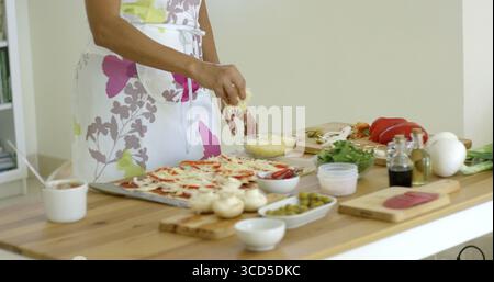 Femme préparant un salami maison et pizza aux champignons dans la cuisine mettant du fromage râpé sur le dessus comme elle se tient à une table en bois vue rapprochée du Banque D'Images