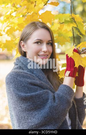 Femme souriante rassemblant des feuilles d'automne dans ses mains portant des moufles rouges posant contre le feuillage d'automne jaune vif Banque D'Images