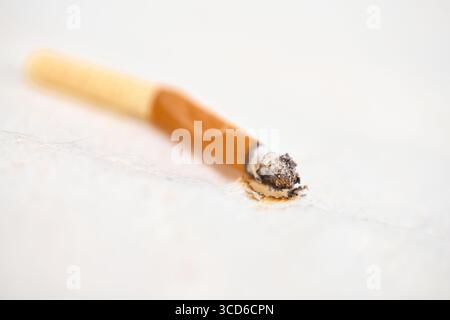 Une cigarette éteinte repose sur une surface texturée claire. Le filtre orange contraste avec la cendre à la pointe. Un éclairage tamisé crée une ambiance calme mais sombre Banque D'Images