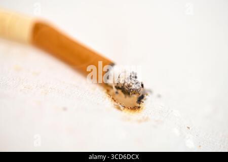 Une cigarette éteinte repose sur une surface texturée claire. Le filtre orange contraste avec la cendre à la pointe. Un éclairage tamisé crée une ambiance calme mais sombre Banque D'Images