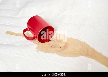 Mug rouge renversé sur tissu texturé blanc, créant une tache étalée. Un éclairage lumineux améliore le contraste entre la tasse vibrante et le fond blanc, emphas Banque D'Images