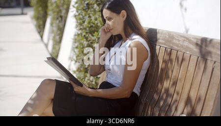 Jolie jeune femme surfant sur Internet sur une tablette comme elle est assise à l'ombre d'un palmier tropical vue de côté Banque D'Images