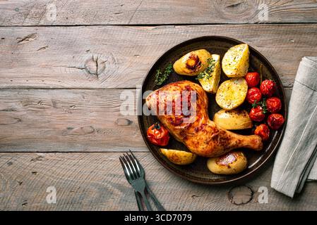 Quartier cuisse de poulet rôti au four avec pommes de terre, tomates et herbes servies sur une table en bois, vue de dessus, espace copie. Repas maison copieux - poulet et ve Banque D'Images