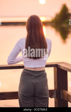 Jeune femme avec de longs cheveux bruns regarde au coucher du soleil sur le lac calme. Des teintes roses et oranges douces remplissent le ciel, créant une ambiance sereine. La terrasse en bois fournit pe Banque D'Images