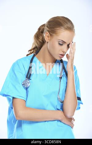 Stressée jeune infirmière ou médecin attirante avec un mal de tête debout dans des gommages bleus avec sa main sur sa tête au-dessus du gris Banque D'Images