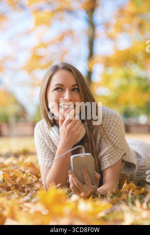 Jolie dame souriante couchée sur des feuilles sèches à Grassy Ground avec téléphone portable. En regardant le cadre du côté droit Banque D'Images