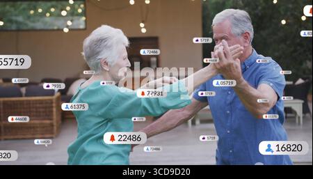 Couple senior dansant portant un chemisier turquoise, chemise bleue sur le patio, avec des icônes sociales flottantes Banque D'Images
