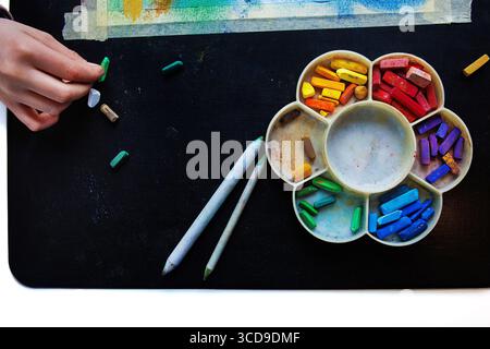 Vue de dessus d'une main arrangeant des craies pastel colorées dans la palette arc-en-ciel avec des outils de mélange sur une table noire Banque D'Images