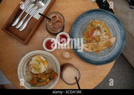 Vue de dessus des pâtes avec pain à l'ail, riz avec poulet frit et oeuf, boissons glacées et condiments servis sur une table de café en bois. Banque D'Images