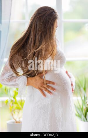 Femme enceinte portant une robe blanche avec les mains sur son ventre à l'intérieur Banque D'Images