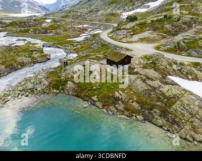 Vue aérienne d'un lac turquoise rencontrant un terrain accidenté et rocheux, une petite structure en bois nichée sur la rive, et une route sinueuse, Hjelledalen, Stryn, Norvège. Banque D'Images