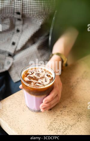 Gros plan de la main d'une femme tenant un verre de café à la lavande glacée dans un café moderne. Infusion froide avec arôme floral, tons pastel doux. Banque D'Images