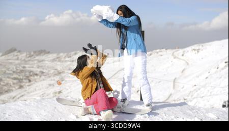Deux jeunes amies jouant dans la neige menaçant de couvrir une avec une grande boule de neige alors qu'elle est assise sur son snowboard dans la neige sur un su de montagne Banque D'Images
