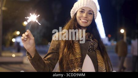 Jeune femme vivante célébrant Noël à l'extérieur lors d'une nuit d'hiver tenant un scintillant dans sa main et riant de la caméra Banque D'Images