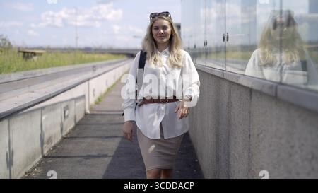 Jeune femme entrepreneure dans des vêtements décontractés intelligents à la mode regardant la caméra et marchant sur le chemin à l'extérieur du bâtiment contemporain, tout en se rendant au travail Banque D'Images