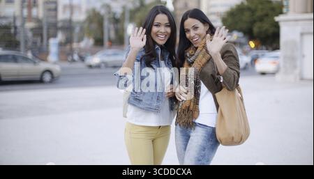 Deux jeunes femmes heureuses à la mode debout dans une rue urbaine agitant à la caméra avec un sourire vif de plaisir Banque D'Images