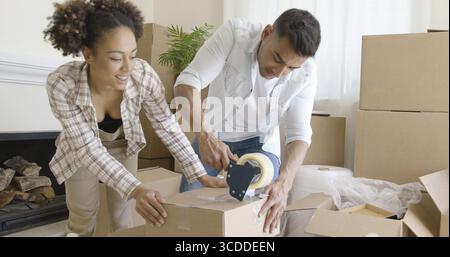 Jeune couple taping cases, comme ils emballer leur maison pour passer à une nouvelle maison à genoux ensemble sur le plancher à travailler en équipe Banque D'Images