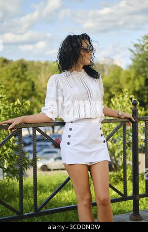 Dans un cadre de parc dynamique, une femme pose en toute confiance dans une blouse blanche et une mini jupe. La lumière du soleil filtre à travers les arbres, mettant en évidence son humanisme détendu Banque D'Images
