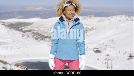 Célibataire belle jeune femme en tenue de ski rose et bleu debout sur la montagne avec un grand sourire tout en regardant sur le côté Banque D'Images