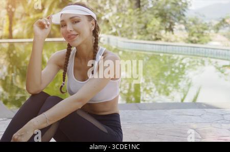 Jeune femme heureuse dans les vêtements de sport touchant la casquette et souriant pour la caméra tout en étant assis près de la piscine pendant l'entraînement en plein air sur la journée ensoleillée sur la station Banque D'Images