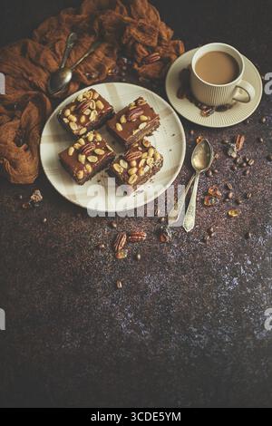 Les morceaux carrés de délicieux gâteau au caramel aux arachides et aux noix du brésil servis avec du café au lait, sur fond rustique. Avec espace de copie, pose à plat Banque D'Images