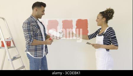 Laughing young couple choisir une couleur de peinture pour rénover leur nouvelle maison debout avec un rouleau et pinceau dans leurs mains en face d'échantillons sur le w Banque D'Images