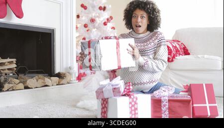 Surpris Jeune femme tenant un grand cadeau de Noël dans ses mains qu'elle s'assoit sur le plancher à la maison présente son ouverture Banque D'Images