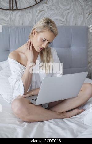 Beatutiful femme dans mens chemise blanche élégante assis dans le lit avec ordinateur portable. Routines matinales Banque D'Images
