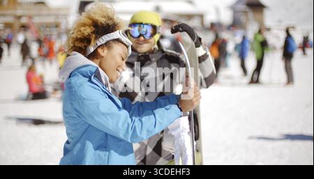 Homme dans des lunettes flirtant avec la femme souriante dans la veste bleue tenant le snowboard sur la piste de ski bondée le jour ensoleillé Banque D'Images