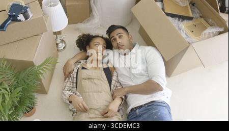 Rêve et heureux jeune couple mixte couché sur le sol parmi des boîtes en carton déballées dans leur nouvel appartement Banque D'Images