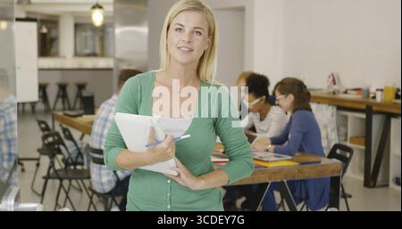 Femme occasionnelle posant avec tas de documents dans les mains et regardant la caméra sur fond de gens dans le bureau Banque D'Images