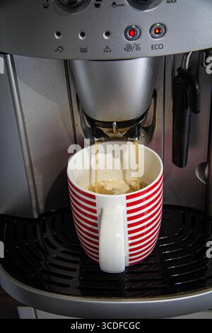 Une machine à café de cuisine domestique créant et versant du café chaud fraîchement préparé à partir de deux becs dans une tasse en porcelaine rouge rayée Banque D'Images