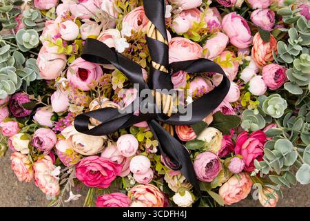 Un bouquet de fleurs artificielles roses, blanches et Bordeaux avec feuillage vert dans un panier avec un ruban de deuil noir sur la poignée, un bouquet funéraire Banque D'Images