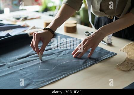 Femme jeune adulte caucasienne marquant le tissu avec la craie de tailleurs sur la table de travail, les mains dessinant des lignes de patron pour la confection de vêtements, des outils de couture et des pièces de patron de papier visibles dans l'espace de travail Banque D'Images