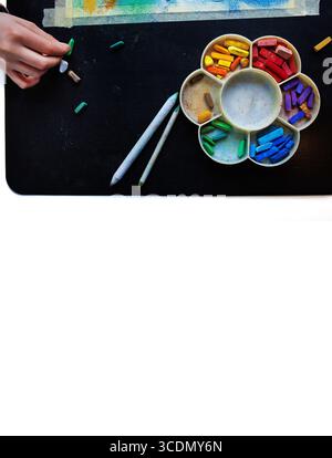 Vue de dessus d'une main arrangeant des craies pastel colorées dans la palette arc-en-ciel avec des outils de mélange sur une table noire Banque D'Images