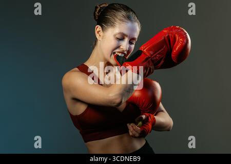 Jeune athlète dans des gants rouges mordant un gant après l'entraînement sur un fond de studio sombre femme intense boxant portrait prêt pour la publicité et l'utilisation éditoriale Banque D'Images