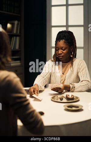 Femme noire d'âge moyen lisant des cartes de Tarot pour le client à la table à l'intérieur Banque D'Images