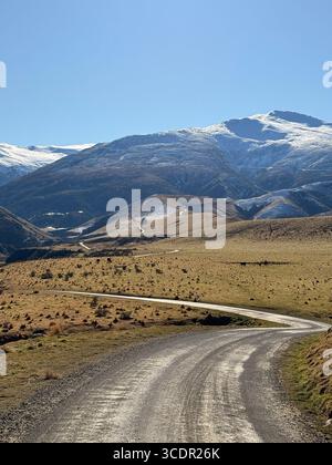 Route de gravier panoramique serpentant à travers des terres agricoles sèches des hautes terres vers des montagnes enneigées sous un ciel bleu clair dans la Nouvelle-Zélande rurale Banque D'Images