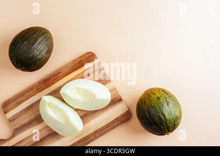 Melon Piel de sapo entier et coupé sur planche à découper en bois sur fond beige. Vue de dessus, pose à plat. Banque D'Images