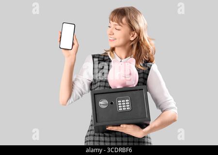 Belle jeune femme heureuse avec coffre-fort, tirelire et téléphone portable sur fond gris Banque D'Images