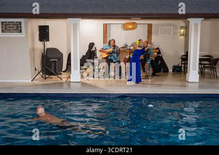 Les personnes qui nagent dans la piscine comme une jeune femme portant un La robe bleue danse une hula hawaïenne à la musique live par deux musiciens hommes matures à l'intérieur de la piscine Banque D'Images