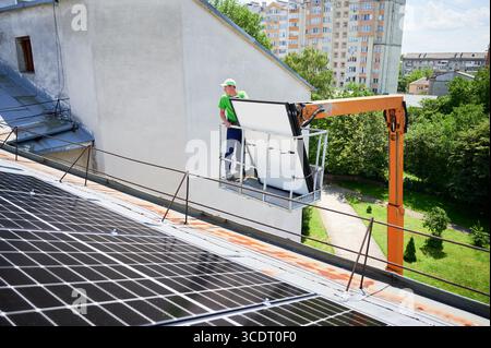 Travailleurs soulevant le panneau solaire photovoltaïque sur le toit métallique de la maison avec l'aide d'un ascenseur de grue. Hommes installateurs installant des modules solaires photovoltaïques à l'extérieur. Concept de génération d'énergie renouvelable. Banque D'Images