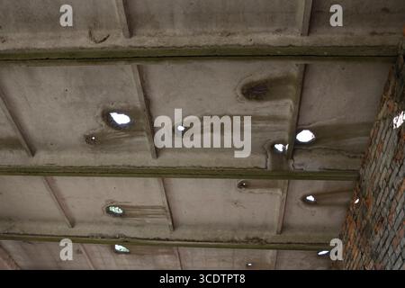 Gros plan d'un plafond en béton endommagé avec plusieurs trous et taches d'eau, avec une partie d'un mur de briques exposé dans un bâtiment industriel abandonné. Banque D'Images