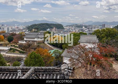 Feuillage d'automne dynamique autour des murs et des toits historiques du château de Himeji, avec vue panoramique sur les gratte-ciel urbain et paysage montagneux Banque D'Images