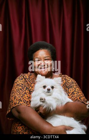 Portrait d'une femme noire d'âge moyen souriant et tenant un chien moelleux dans la cabine photo, femme regardant directement la caméra, chien faisant face à l'avant avec une expression détendue Banque D'Images