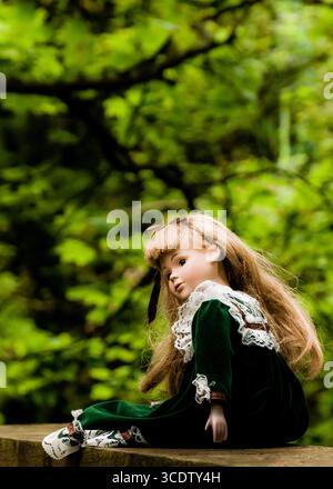Une poupée en porcelaine avec de longs cheveux blonds et une robe verte se trouve sur une surface en bois, avec un fond vert flou de feuillage. Banque D'Images