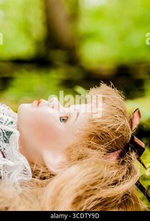 Gros plan du visage d'une poupée en porcelaine, couchée sur un lit de feuillage vert. La poupée a les cheveux blonds, les yeux bleus, et un arc rouge dans ses cheveux. Banque D'Images