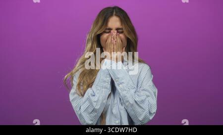 Jeune femme blonde grimace visiblement et tient les deux mains sur les joues dans un studio photo violet ; douleur angoisse détresse. Banque D'Images
