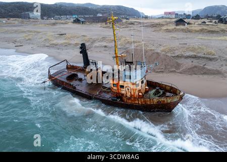 Vue aérienne d'un naufrage rouillé échoué sur une plage de sable, les vagues s'écrasant doucement contre sa coque altérée sur fond d'un village côtier isolé, Teriberka, oblast de Mourmansk, Russie. Banque D'Images